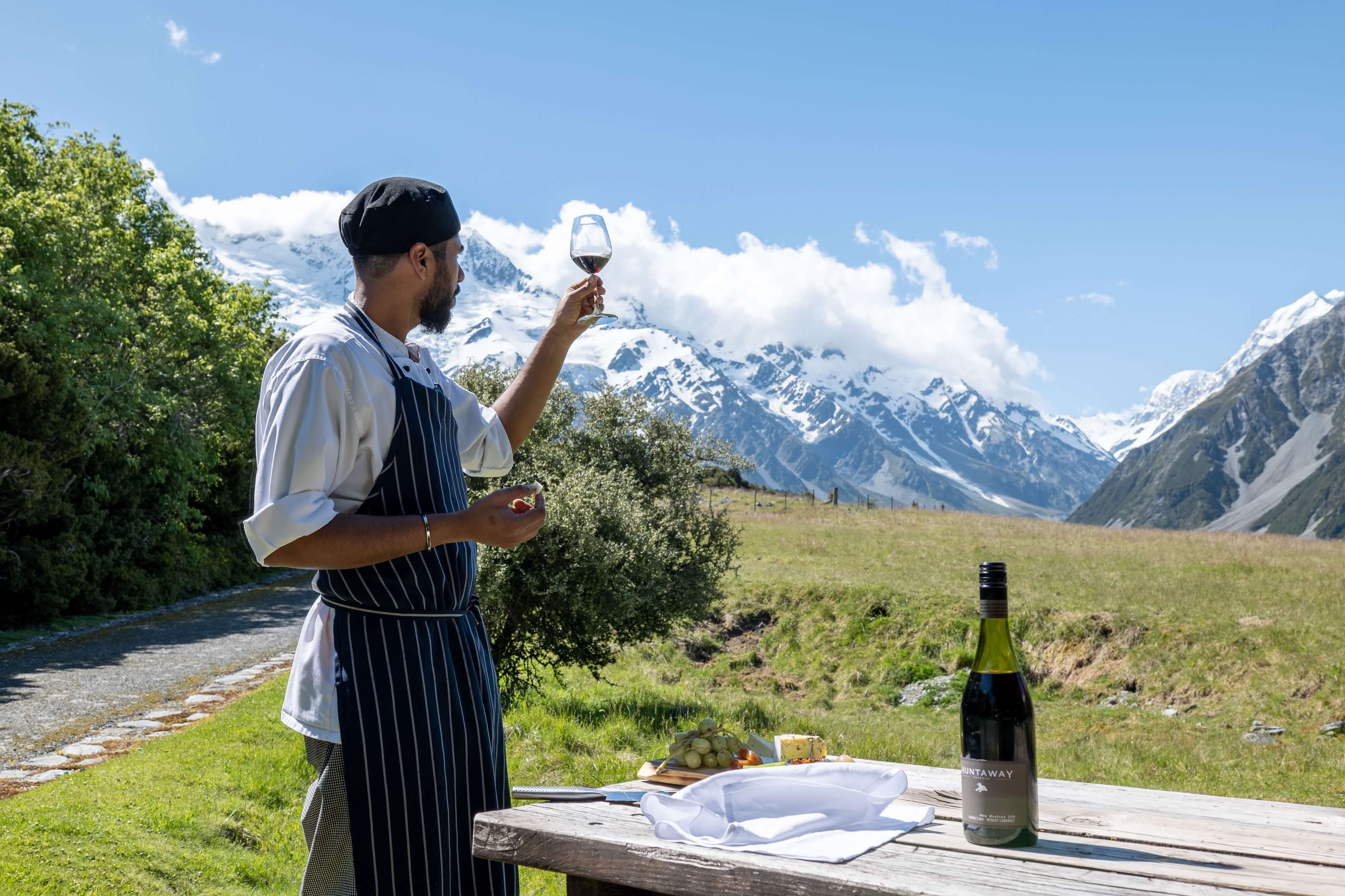 Chef in apron holding red wine glass outdoors with mountains, field, and wine bottle on wooden table.