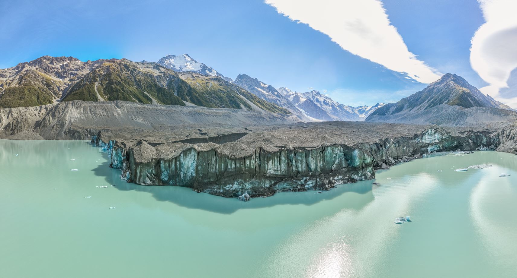 Glacier with icy cliff edges flowing into milky blue water, surrounded by rocky mountains under blue sky.