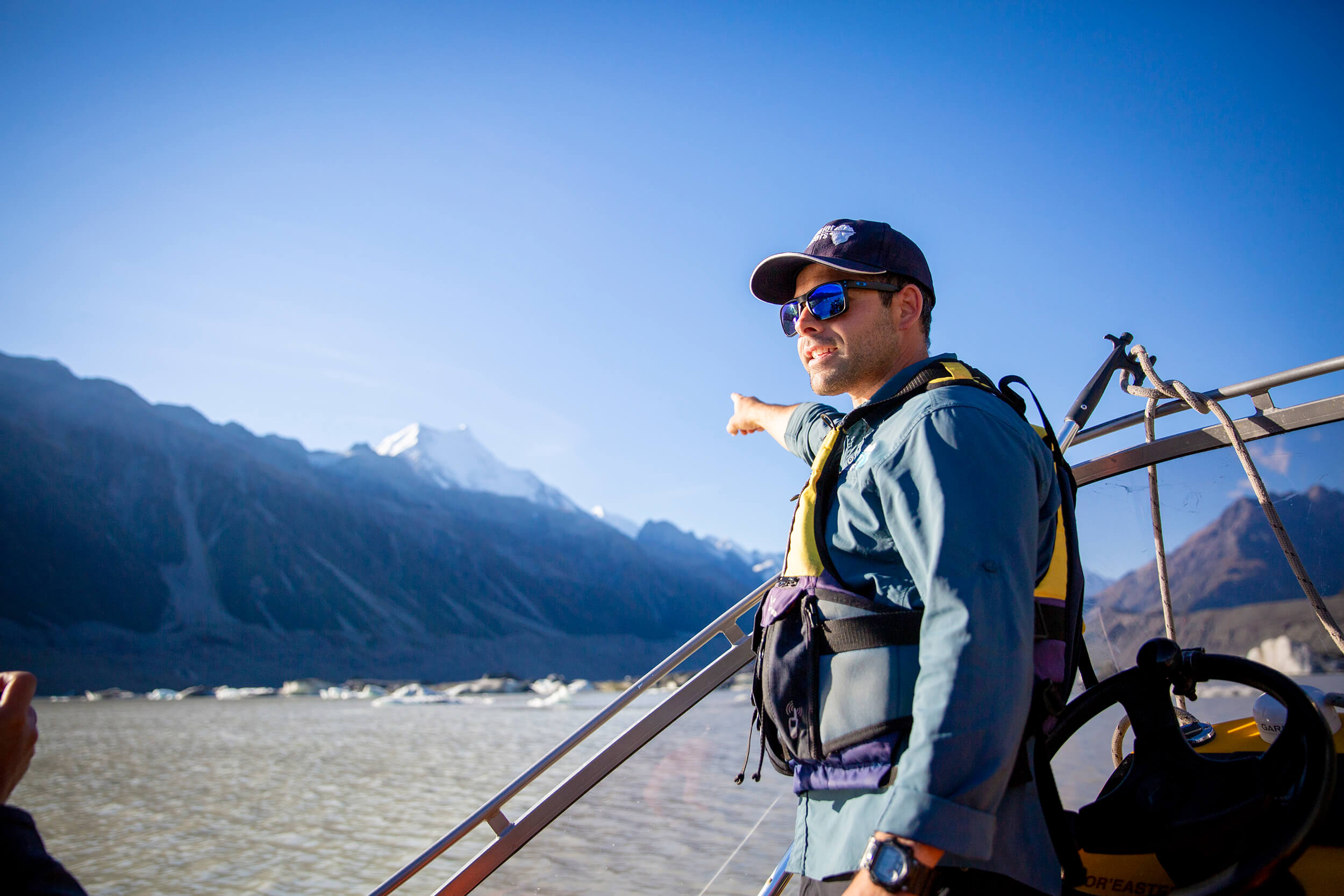 Man wearing sunglasses and life vest points toward snow-capped mountains from a boat on a glacier lake.
