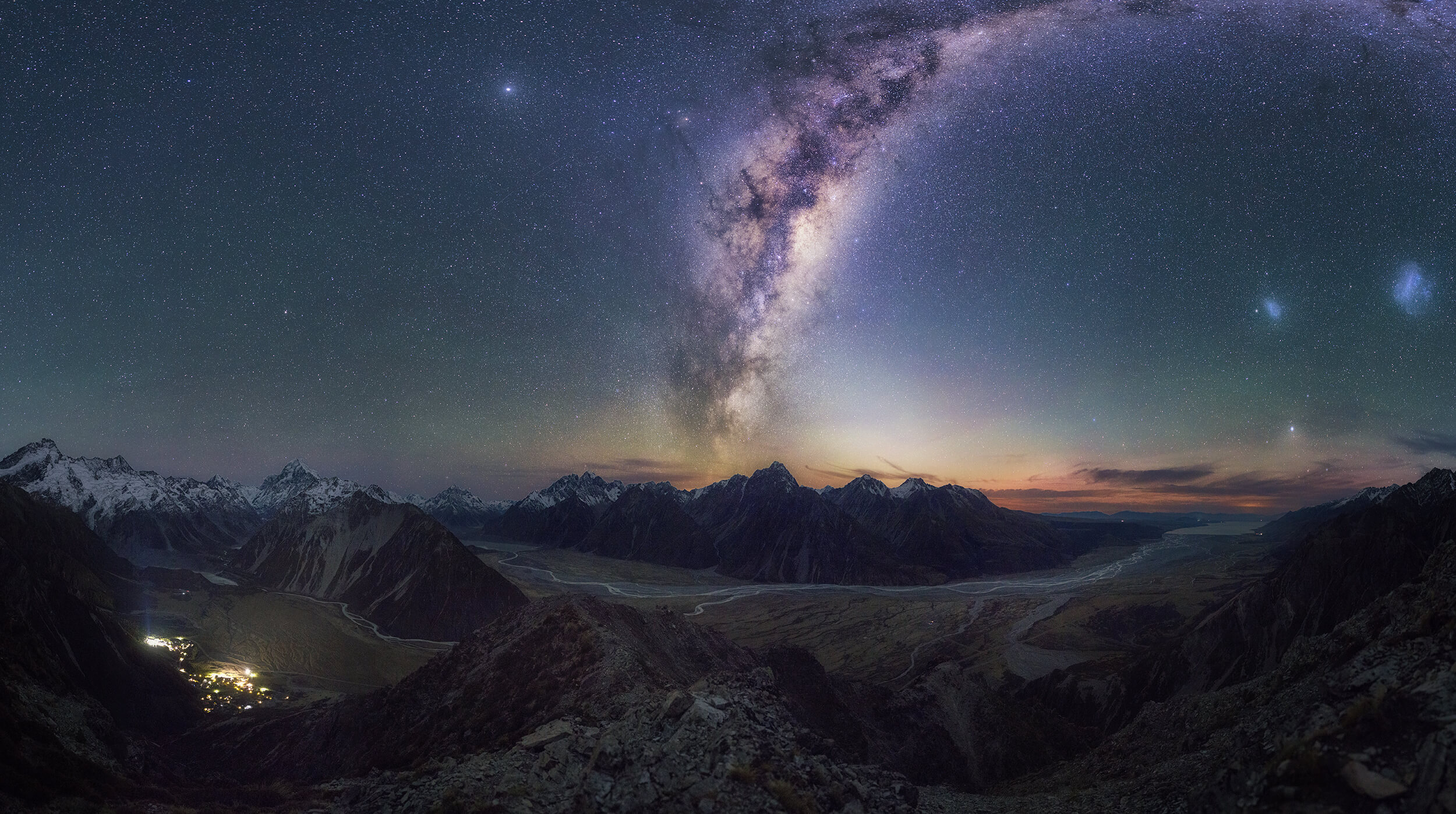 Milky Way galaxy arcing over a mountain range with a starry night sky and a small lit village below.
