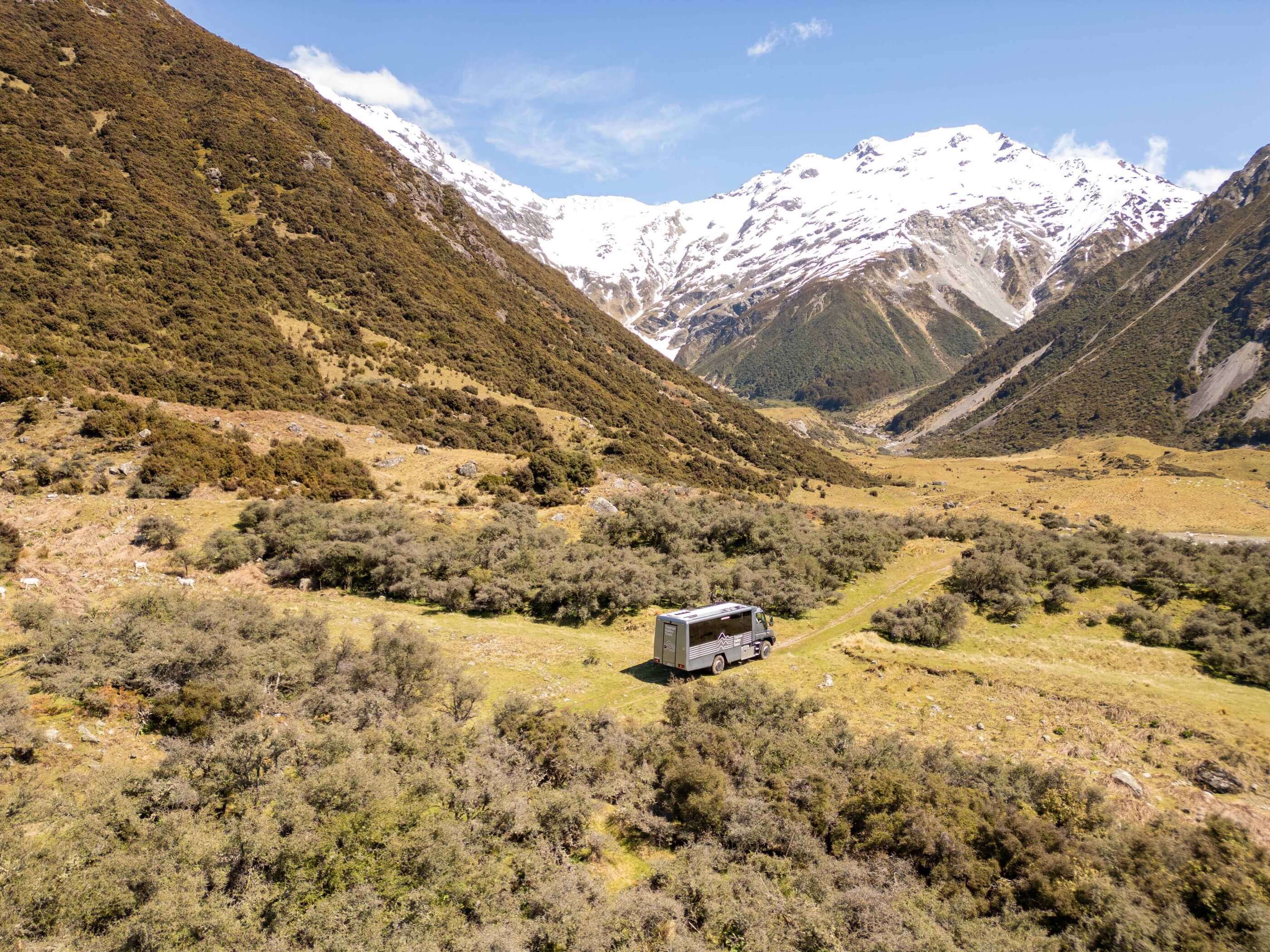 Tour bus parked on grassy terrain surrounded by hills and snow-capped mountains in the background.