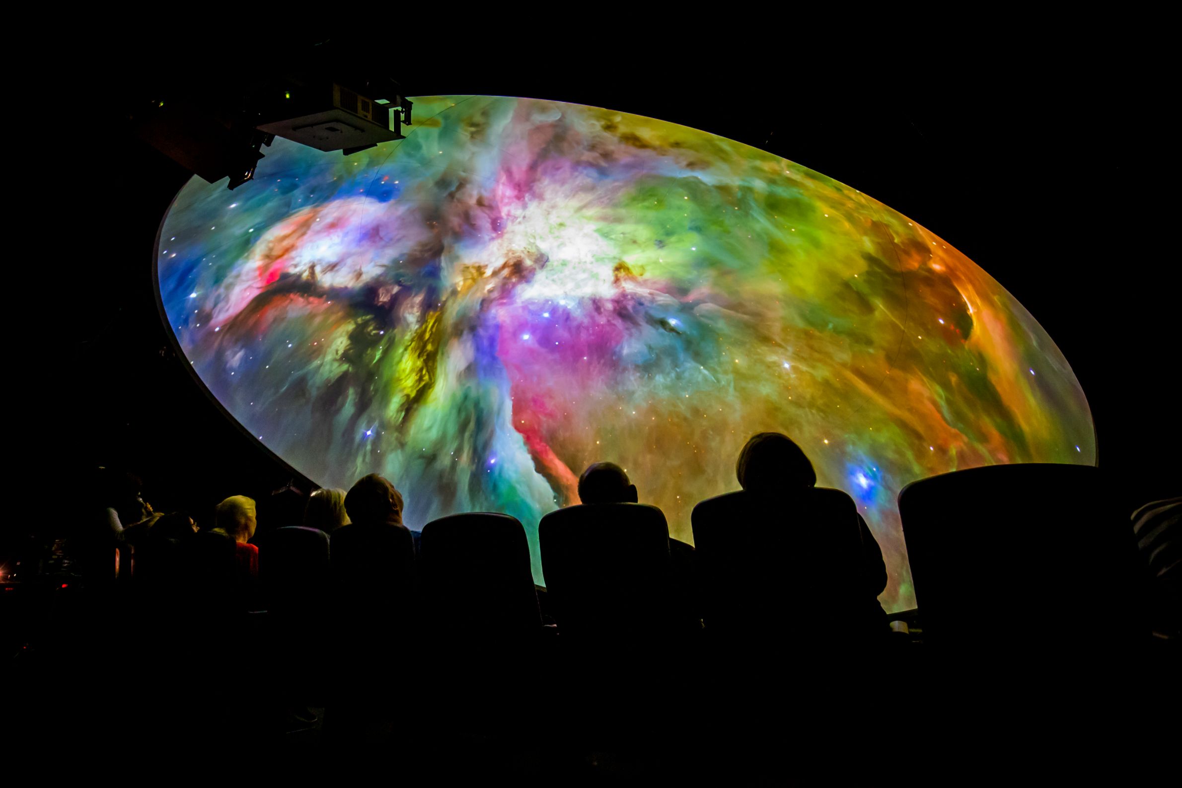 People seated in a dark planetarium watching a colorful nebula projection on a dome ceiling.