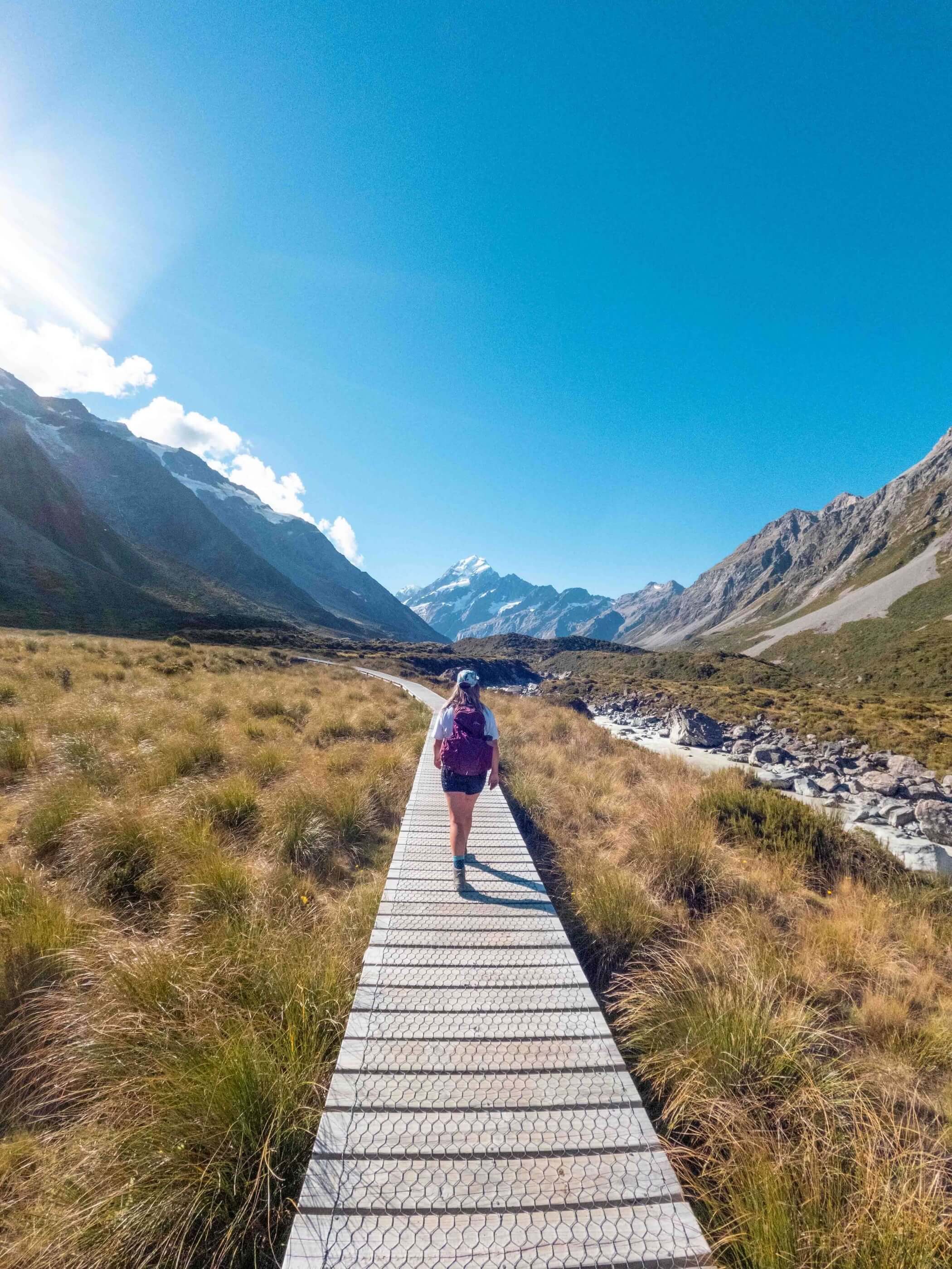 Person hiking on a wooden path through a valley with mountains and a river under a clear blue sky.