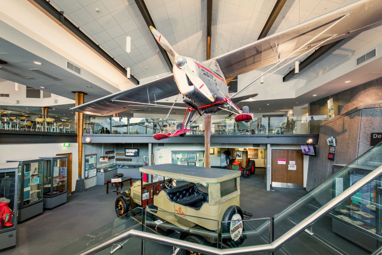 Vintage car below a hanging vintage airplane inside a modern museum with glass railings and displays.