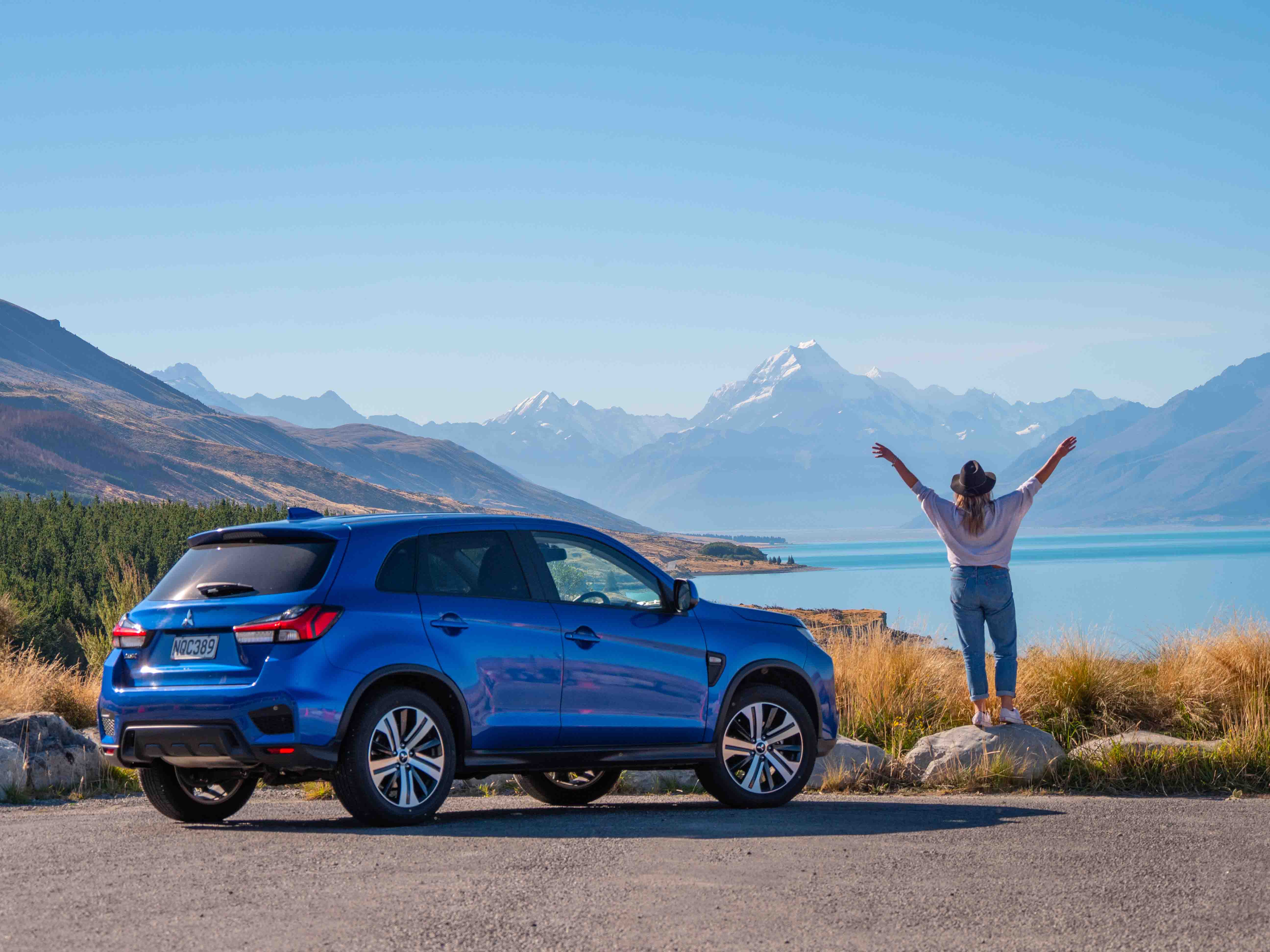Blue SUV parked by a lake with a person raising arms facing snow-capped mountains under clear sky.