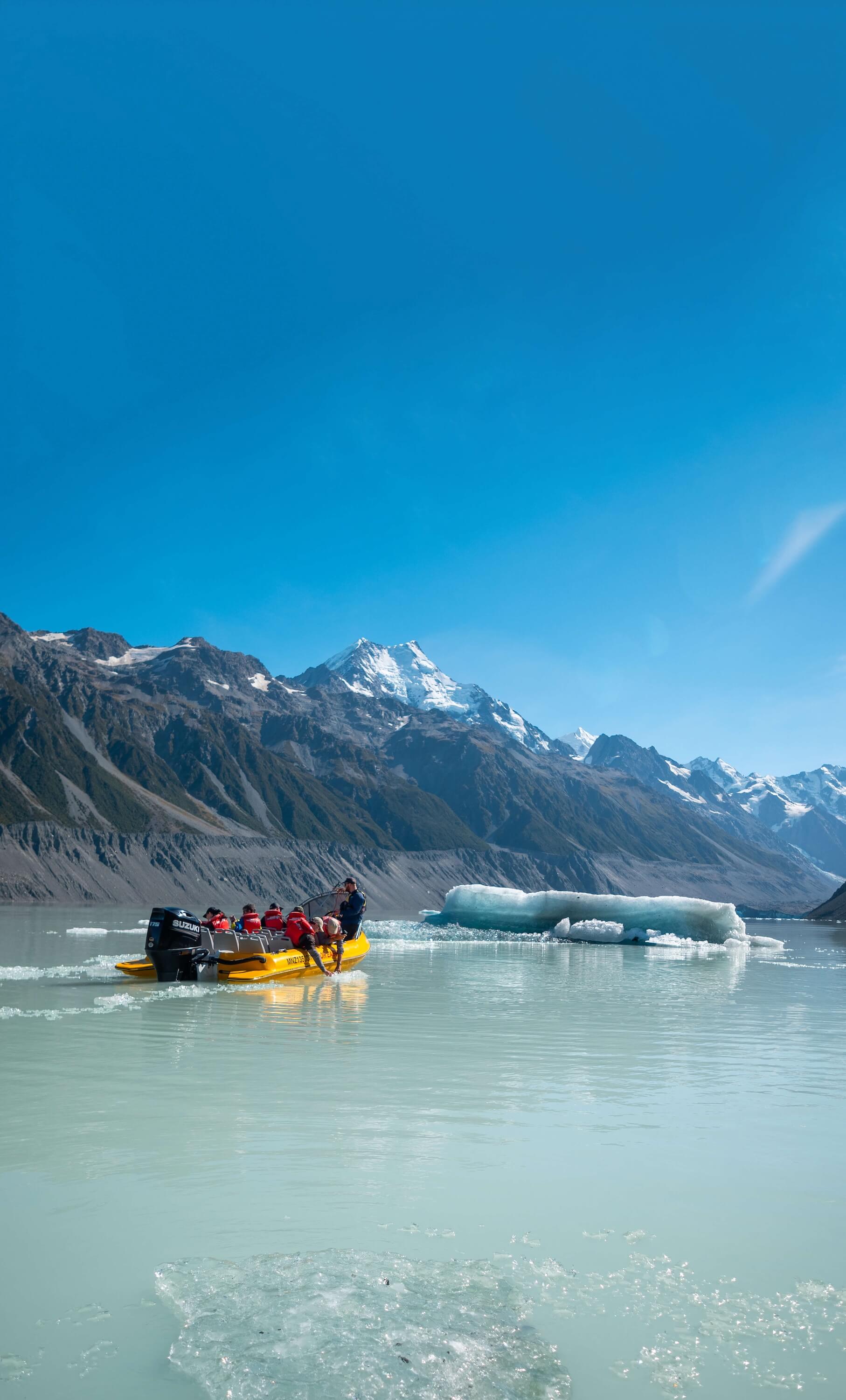 People in red jackets riding a yellow motorboat on icy lake with snow-capped mountains in the background.