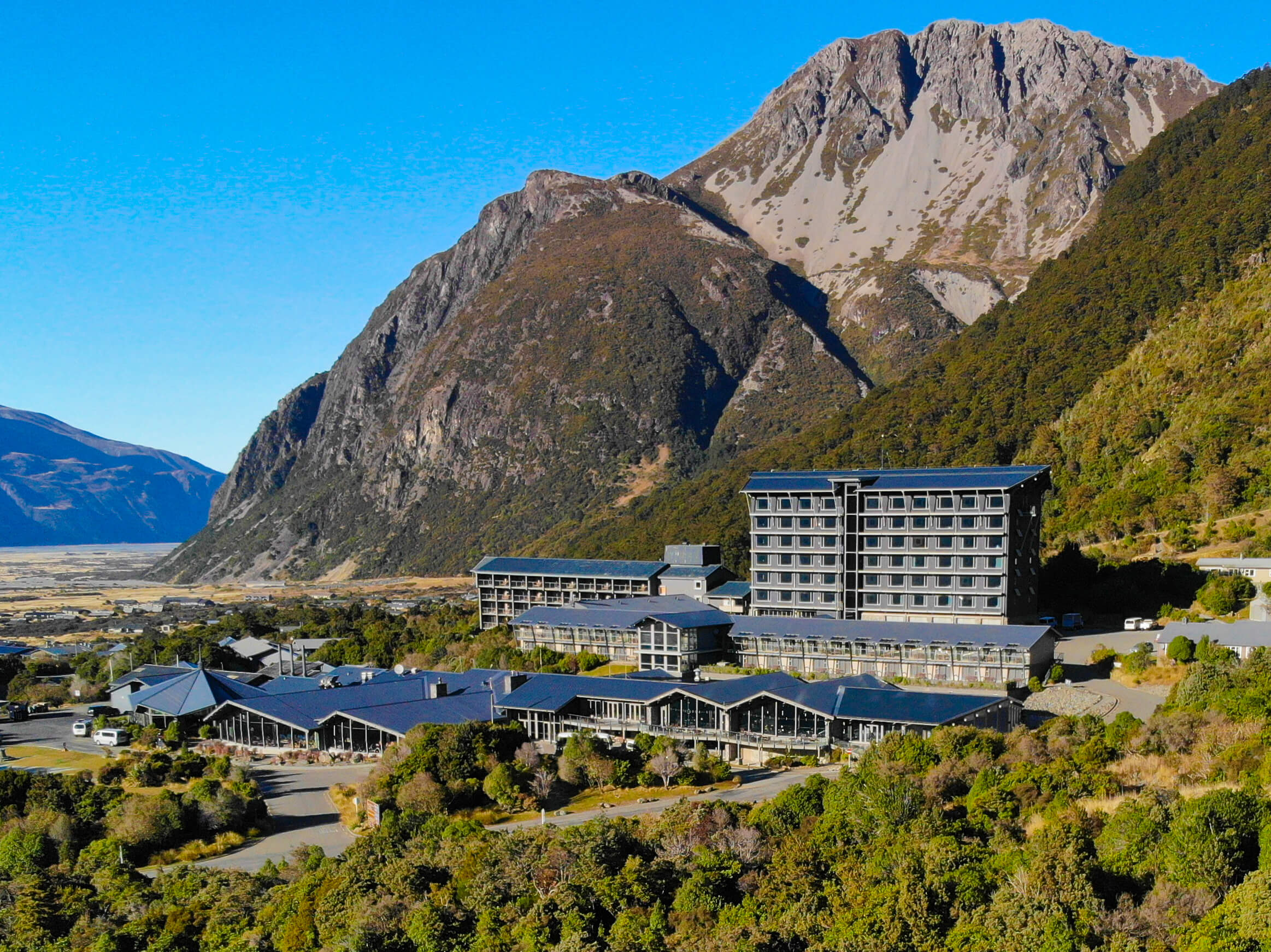 Large hotel complex nestled among green hills with tall rocky mountains and clear blue sky behind.