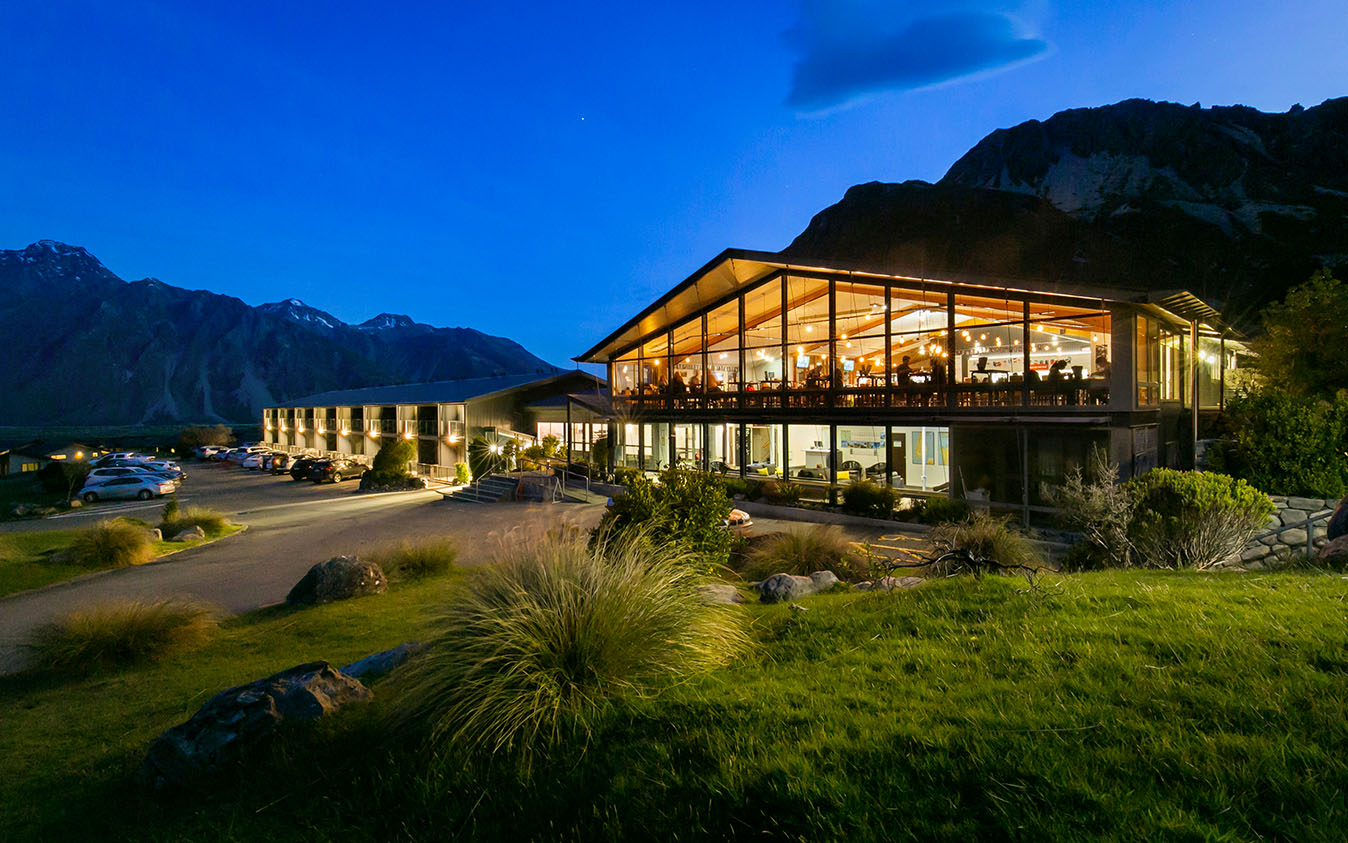 Modern hotel building at dusk with large lit dining area and mountain backdrop.