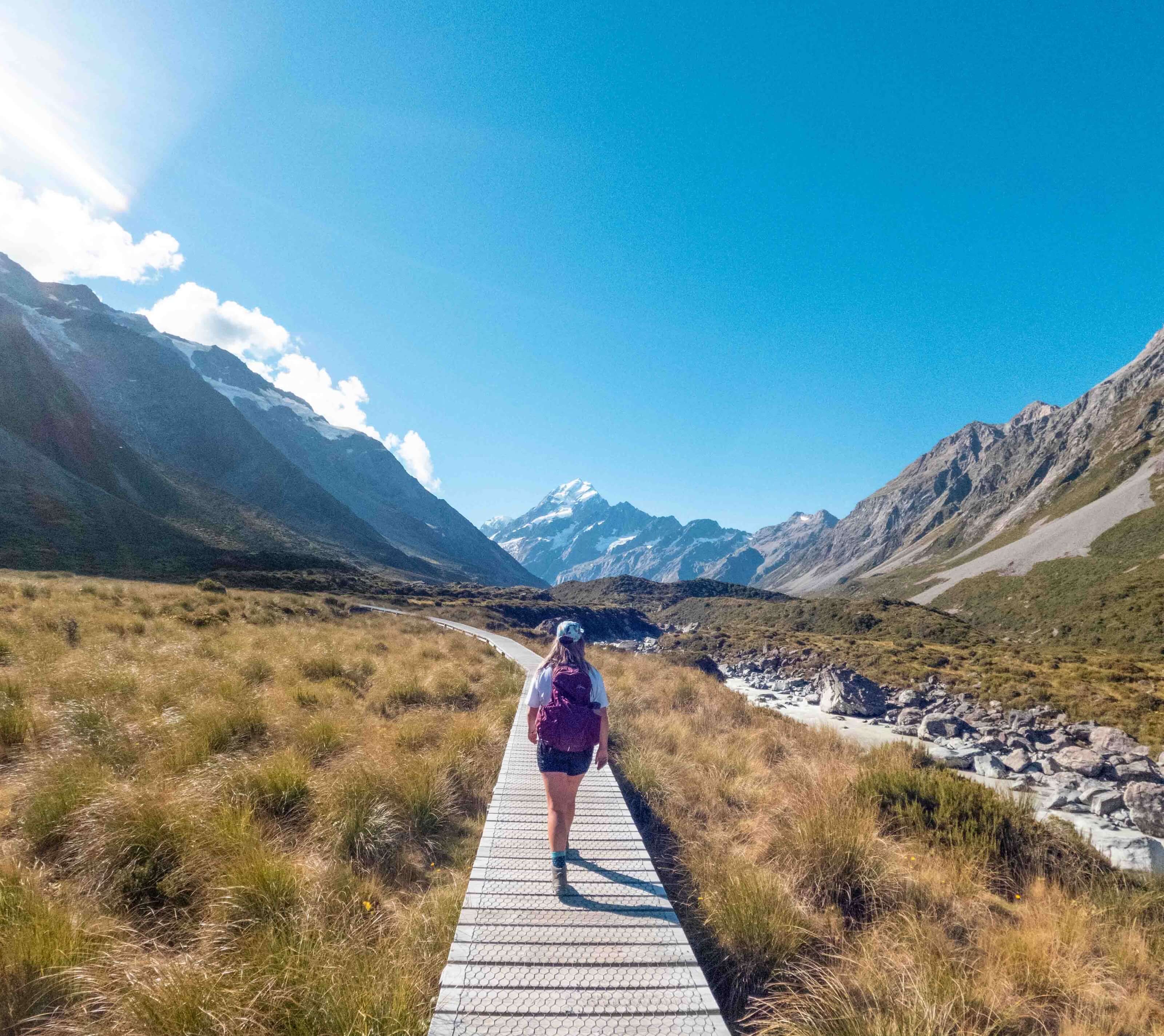 Person with a backpack walking on a boardwalk trail through a mountainous landscape under a clear blue sky.