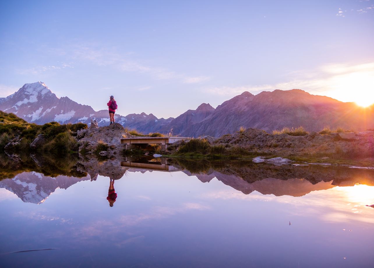 Person in pink jacket standing on rock by a mountain lake during sunset with clear mountain reflections.