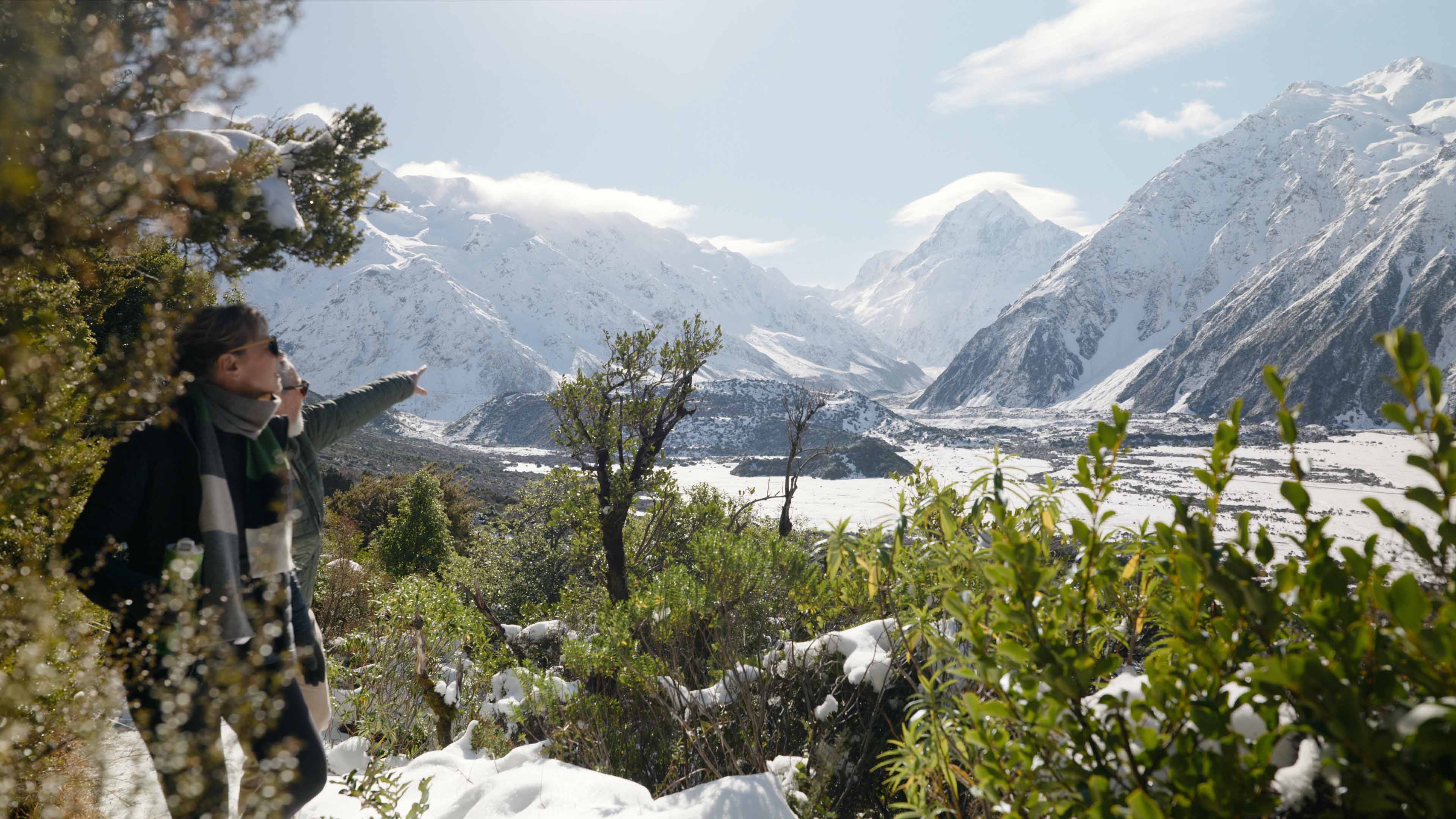 Two hikers with sunglasses in snowy mountain landscape with bushes and clear sky.