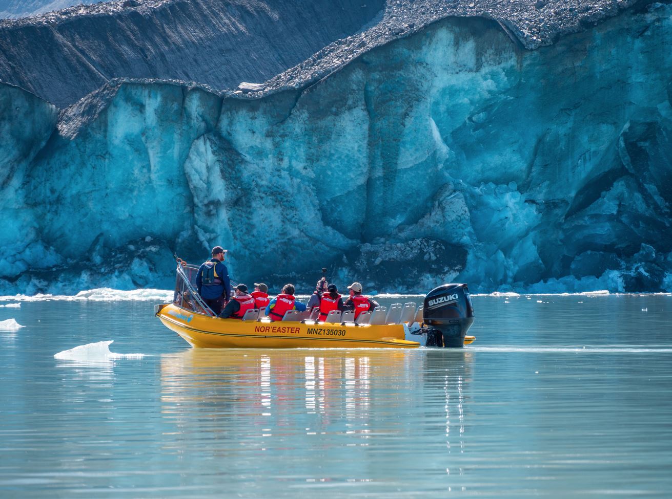 Yellow motorboat with people wearing red life jackets near a large glacier on calm water.