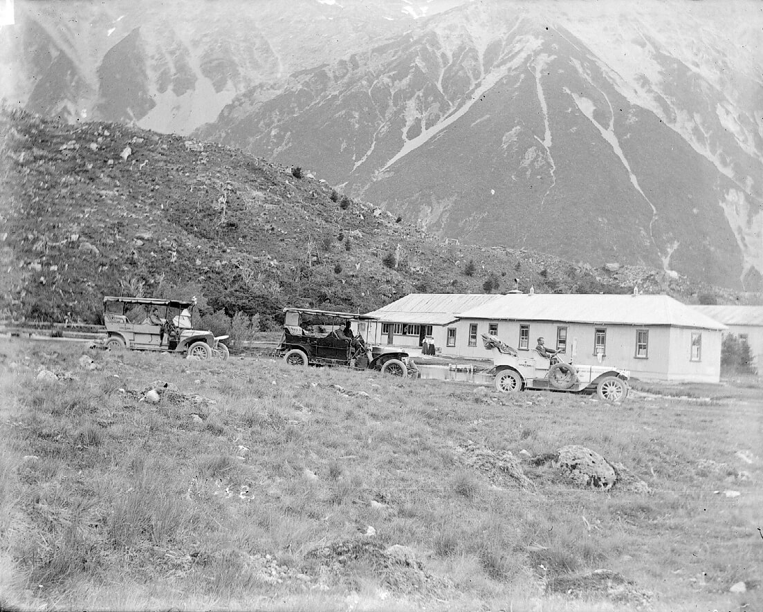 Three vintage cars parked on rocky grassland with a building and mountains in the background.