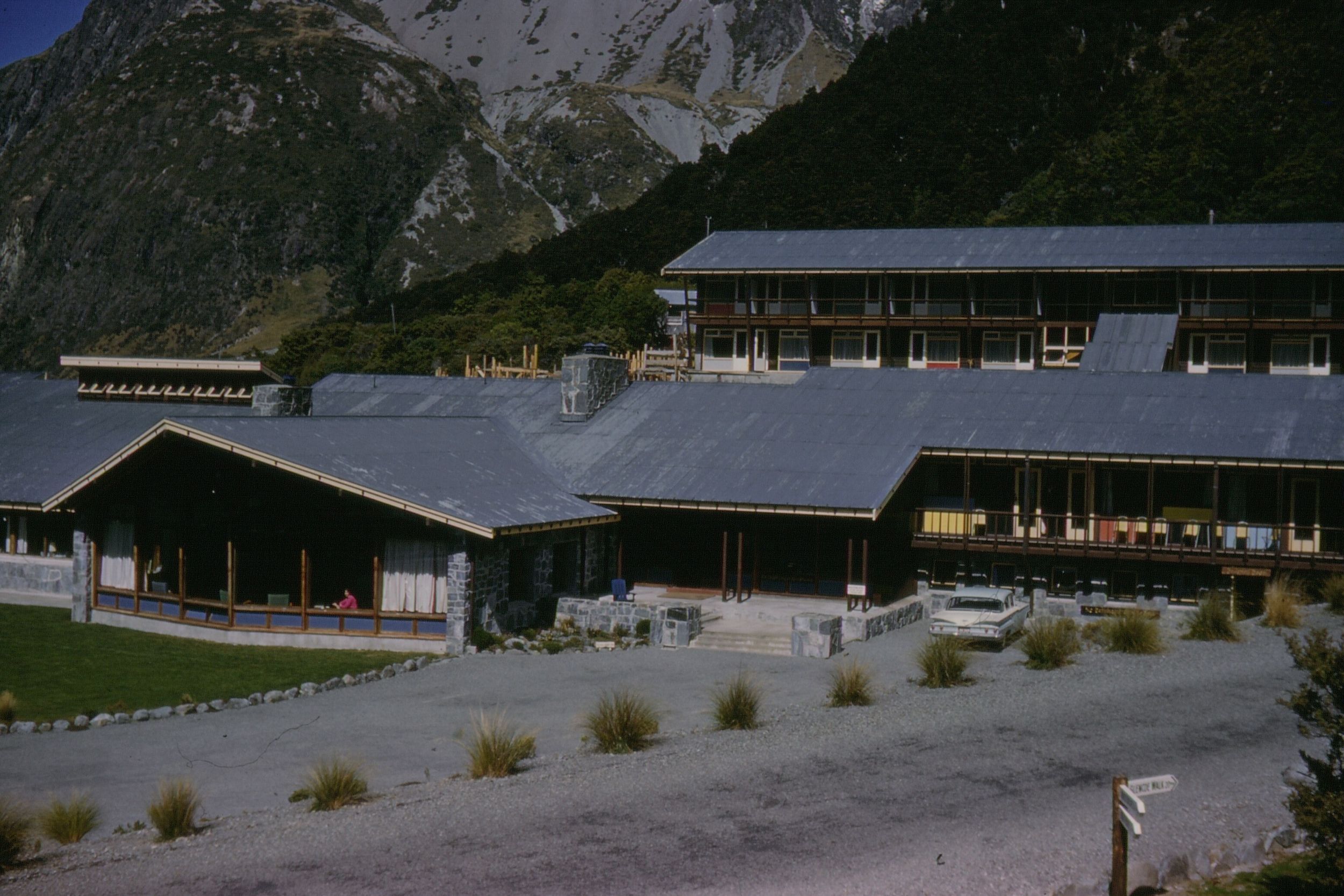 Large lodge with gray roofs set against steep forested mountains under a clear sky.