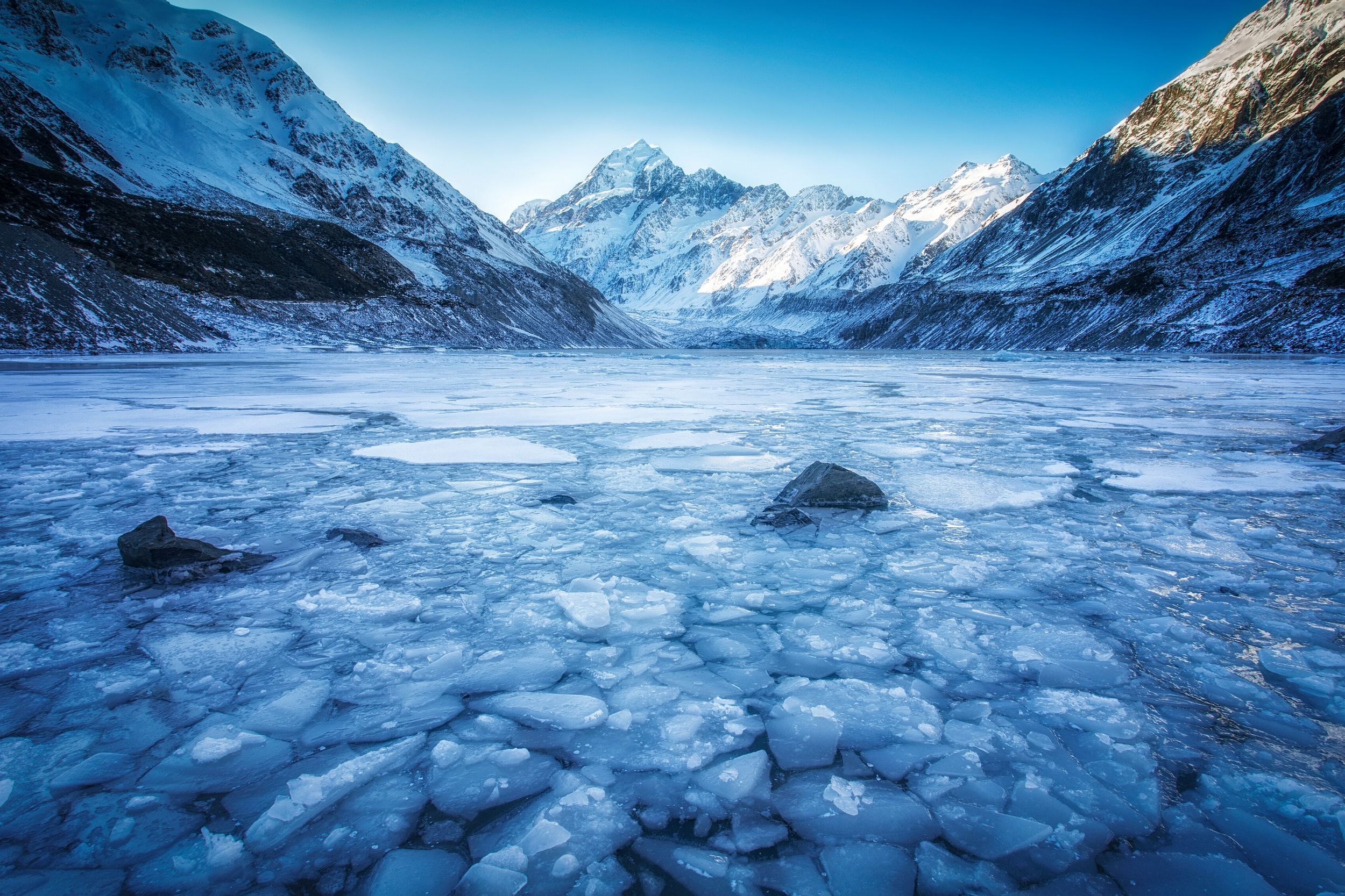 Frozen lake with broken ice pieces surrounded by snow-covered mountain peaks under a clear blue sky.