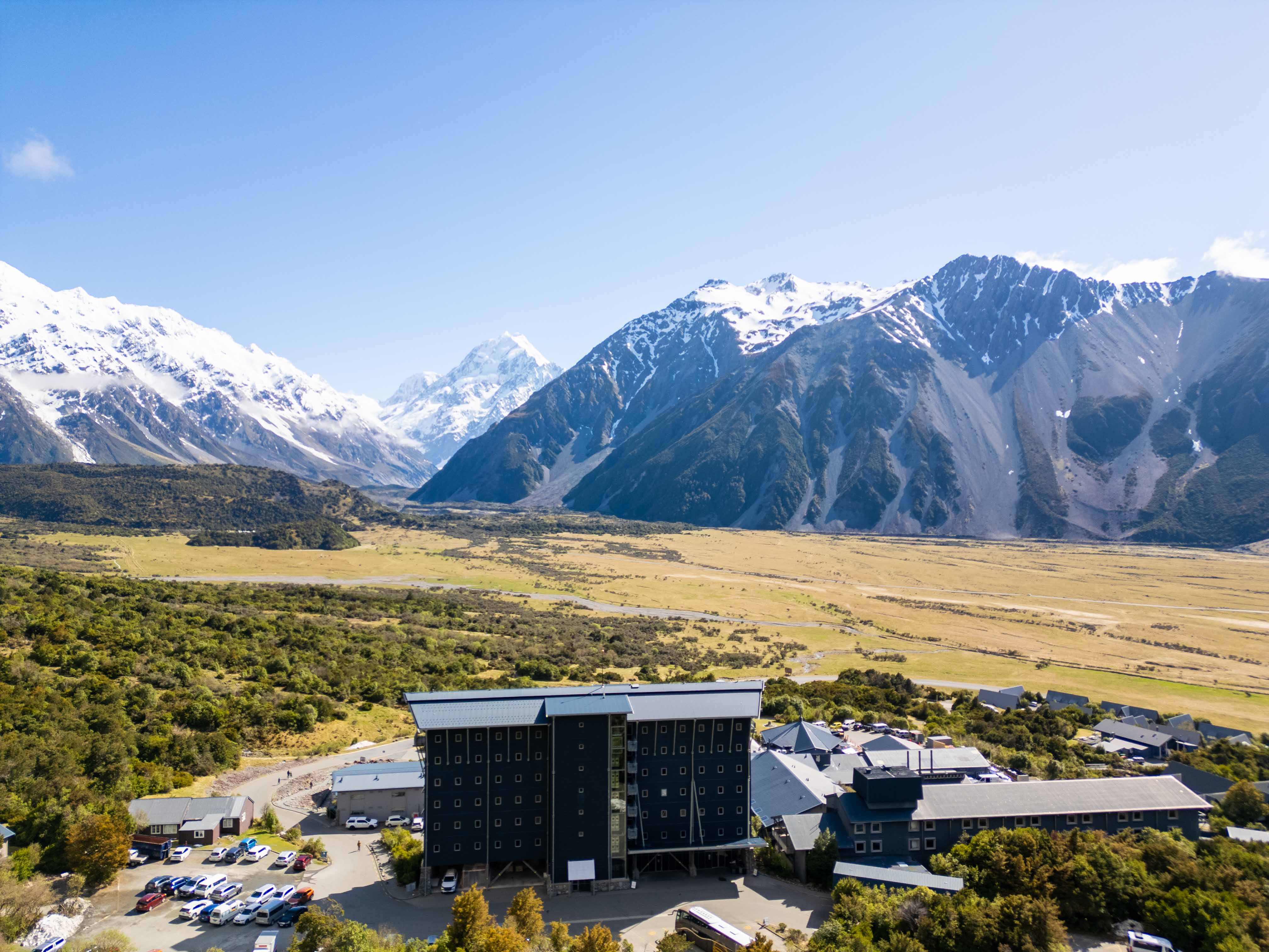Hotel building near lush greenery with snow-capped mountains under a blue sky in the background.