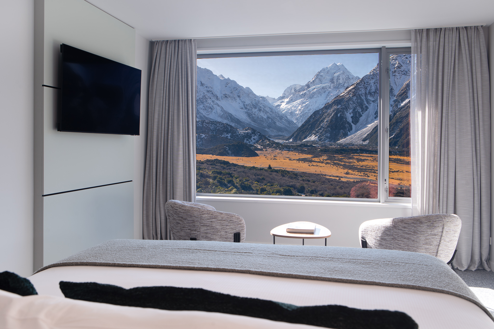 Modern bedroom with gray chairs and table by window showing snow-capped mountains and valley view.