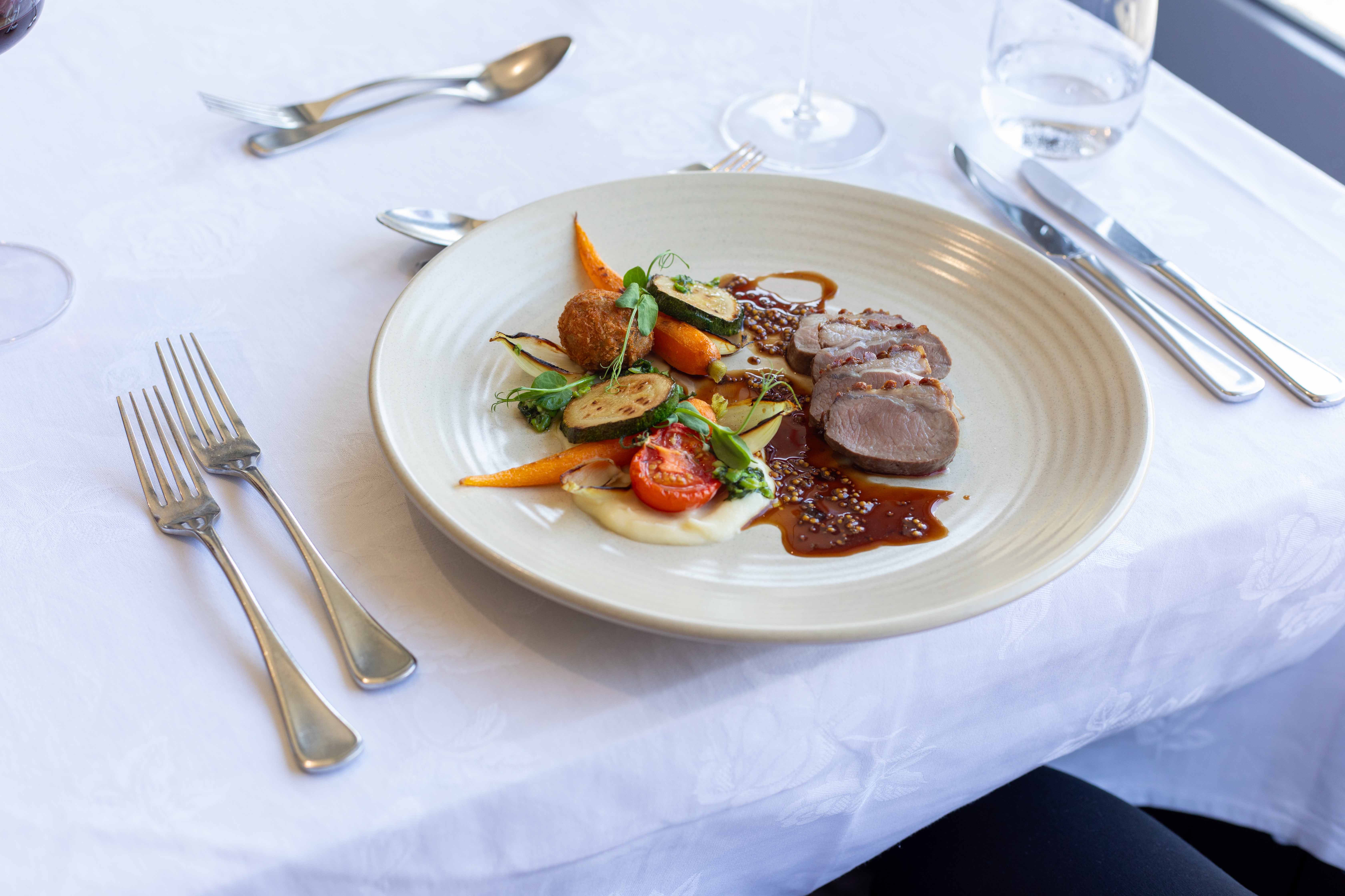 Plate with sliced meat, carrots, grilled zucchini, cherry tomato, greens, and sauce on white tablecloth.