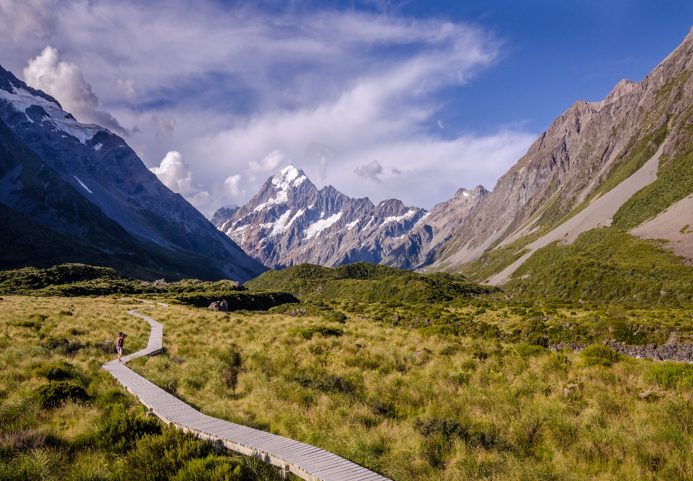 Person walking on wooden path through grass valley with snow-capped mountains under blue sky.