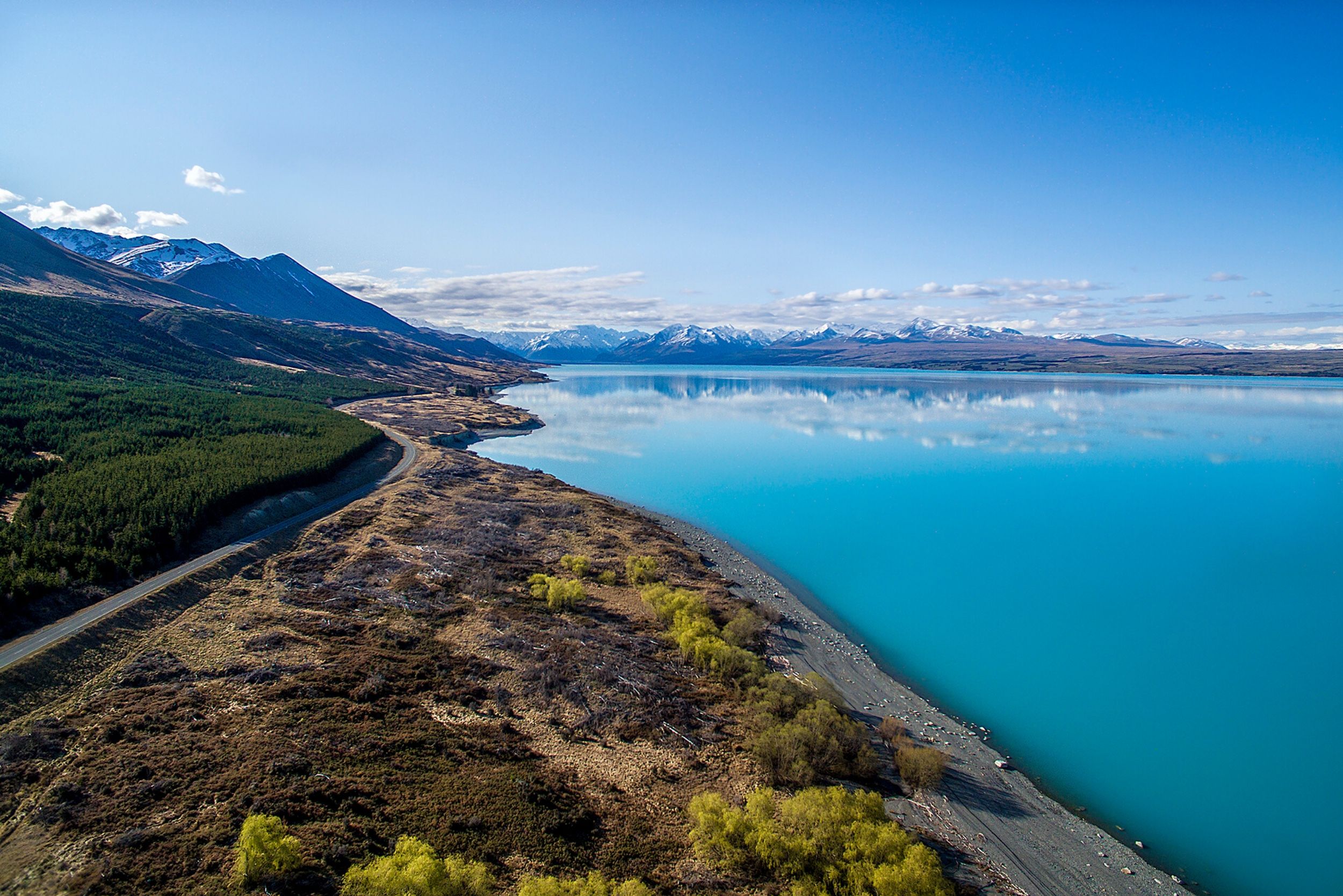 Calm blue lake with mountain range and clear sky reflected, bordered by road and vegetated land.